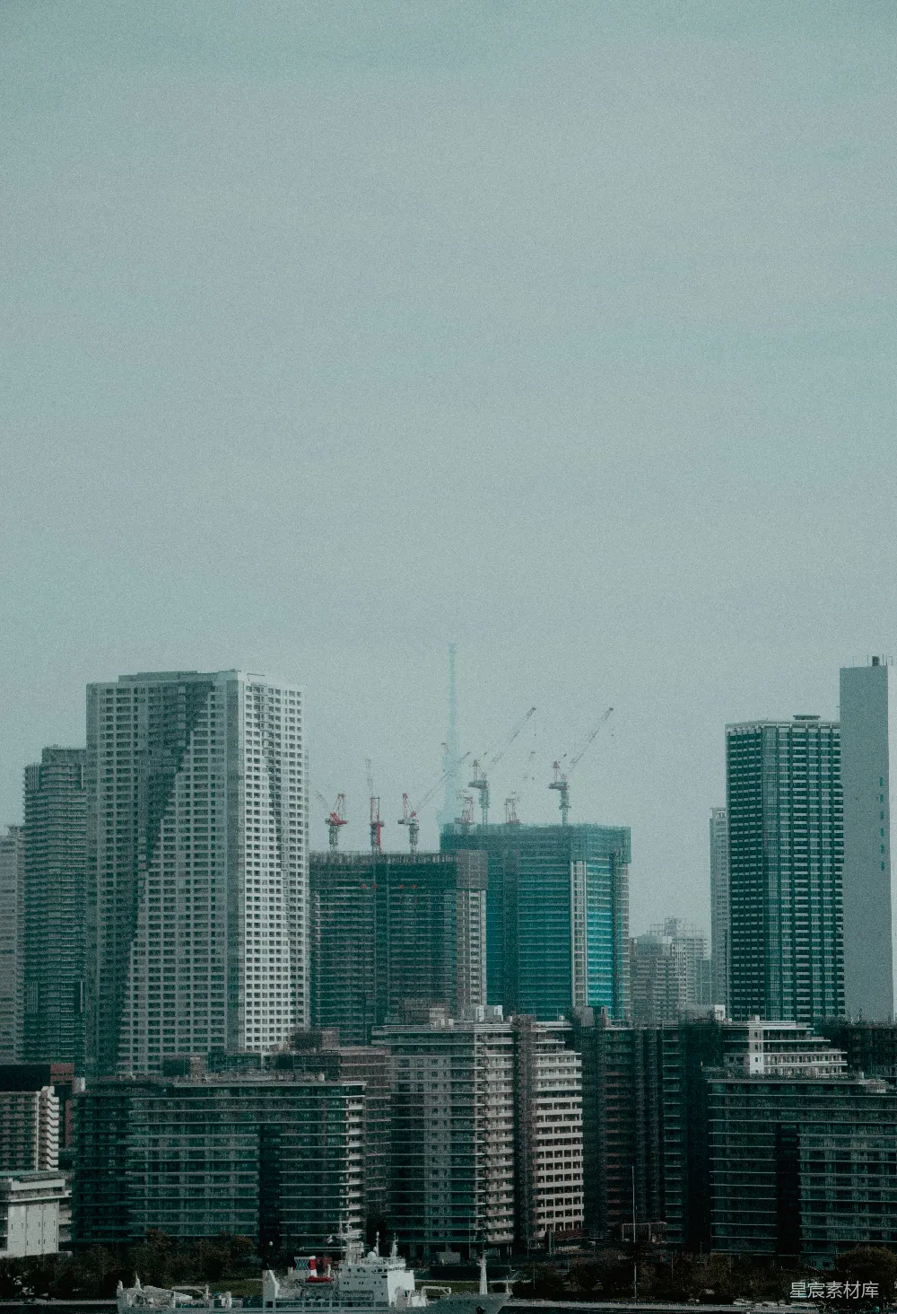 这张图片展示了城市的远景，高楼大厦与正在建设中的建筑形成对比。天空中飘着雾气，给整个场景增添了一丝朦胧感。远处的起重机表明这里正在进行大型建筑项目的施工。整体色调偏冷，给人一种宁静而忙碌的感觉，适合用作都市生活的背景或工业题材的视觉素材。