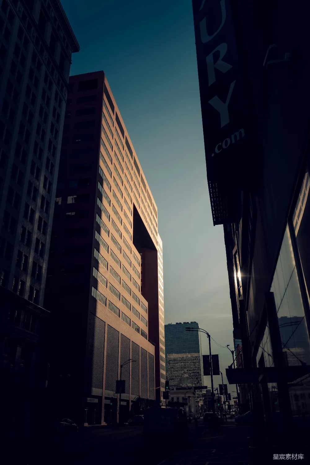 这张图片展示了一个城市的黄昏街景，高楼大厦在夕阳的余晖中显得格外壮观。画面色调偏暗，天空中云层稀疏，给人一种宁静而略带忧郁的感觉。街道上没有行人和车辆，营造出一种静谧的氛围。适合用作城市风光或日落主题的背景素材。