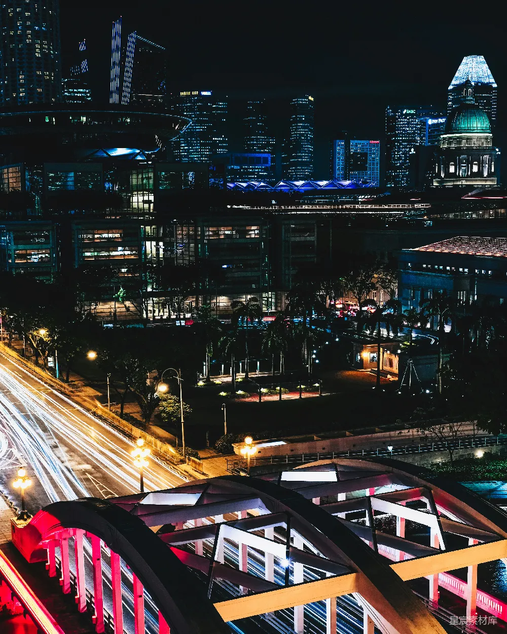 这张图片展示了新加坡的夜景，现代化的摩天大楼灯火辉煌，灯光璀璨，形成一幅美丽的城市天际线。前景中的红色建筑结构与街道上的光轨交相辉映，营造出一种动感与静谧并存的氛围。适合用于旅游宣传、城市风光展示或现代都市的主题设计。