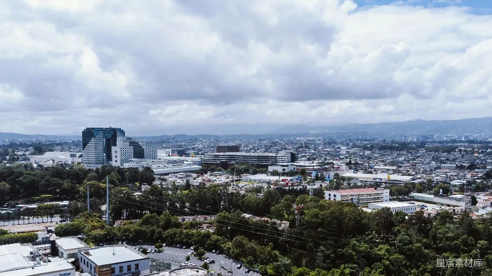 这张图片展示了城市的全景俯瞰，天空中布满了大片的云彩，下方是密集的建筑和道路，远处是连绵的山脉和绿色的树木。整体风格清新自然，给人一种宁静而充满活力的感觉。适合用于城市规划、建筑设计和自然环境相关的展示。