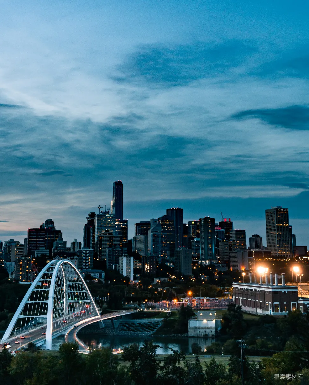 这张图片展示了城市的夜景，天空中布满了稀疏的云彩，地平线上是现代化的高楼大厦，中间有一座白色的桥梁连接着城市和河岸，桥下是河流。远处的灯光照亮了城市，给人一种宁静而充满活力的感觉。适合用作城市风光、旅游宣传或城市生活主题的背景图片。