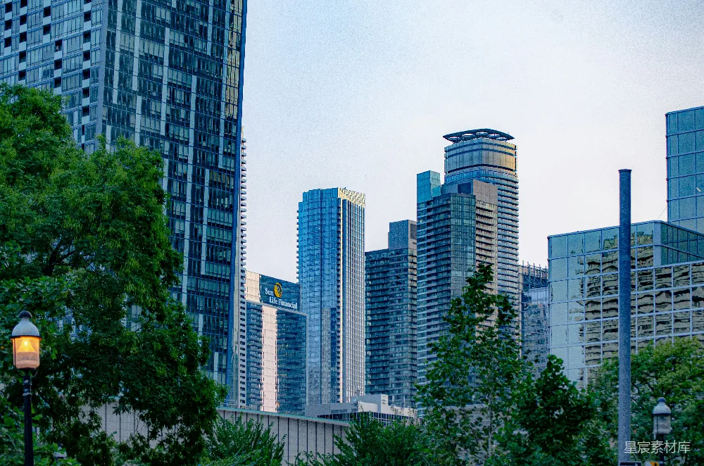 这张图片展示了一座现代化城市的天际线，高楼大厦林立，玻璃幕墙反射出蓝天白云。前景中有绿树和路灯，营造出一种自然与都市结合的和谐氛围。适合用于城市风景、建筑设计或都市生活相关的主题。
