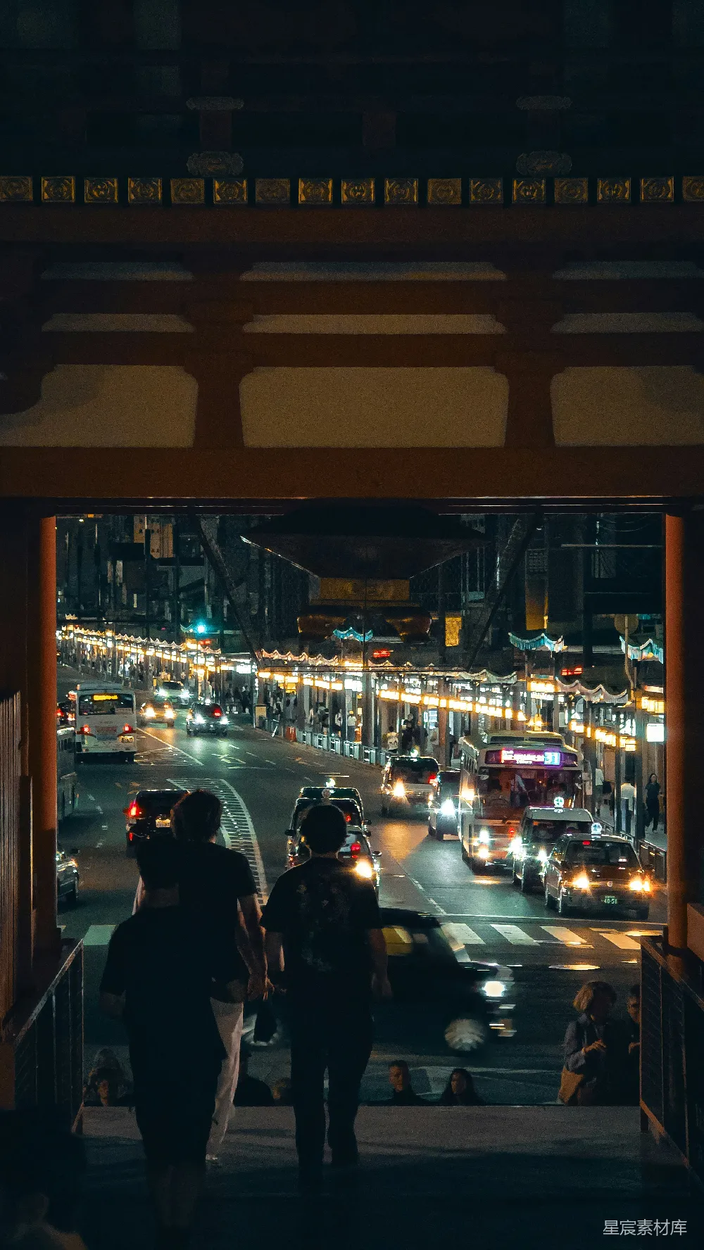 这张图片展示了一个夜晚的城市景象，从一个传统建筑的门廊中窥视出去。街道两旁是繁忙的车流和行人，远处的灯光勾勒出城市的轮廓，营造出一种现代与传统交融的氛围。适合用作城市夜景、旅行目的地或都市生活的视觉素材。