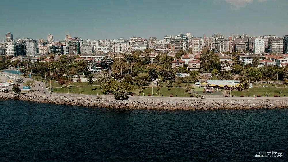 这张图片展示了一座海滨城市的全景，背景是密集的高层建筑，前景则是宁静的海岸线和延伸到海中的岩石堤坝。海岸边有一片绿意盎然的公园和几处休闲场所，阳光明媚，树木和建筑物在蓝天的映衬下显得格外清晰。适合用于城市规划、旅游宣传或房地产广告等场景。