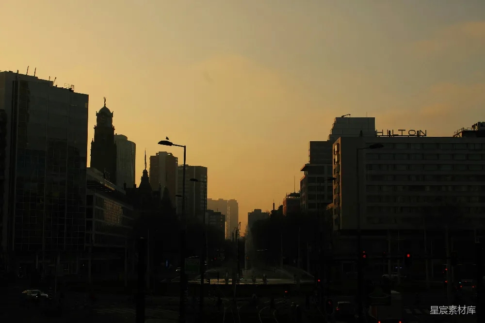 这张图片展示了黄昏时分的城市剪影，天空呈现出温暖的橙黄色调，建筑物在落日余晖下形成鲜明的轮廓。街道两旁的高楼大厦和历史建筑在夕阳的照耀下显得轮廓分明，给人一种静谧而庄重的感觉。适合用作城市风光、城市剪影或黄昏美景的背景。