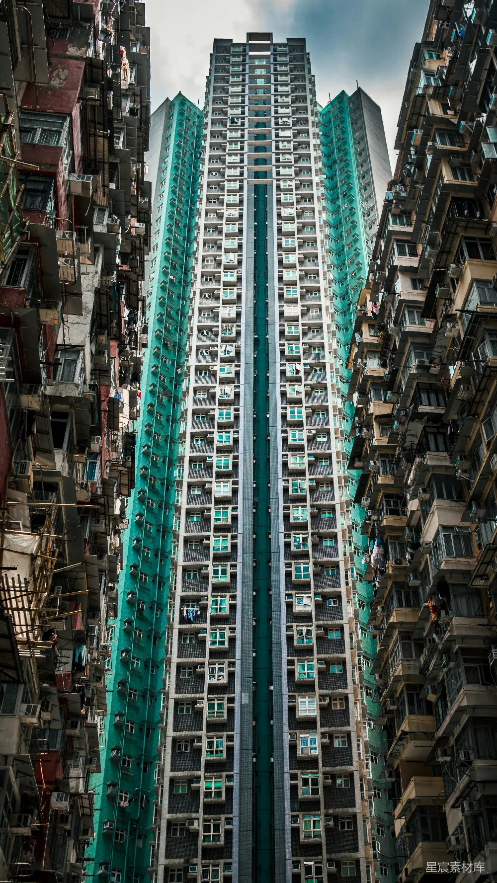 这张图片展示了香港的一座摩天大楼，其独特的绿色外墙在高楼林立的环境中显得格外醒目。大楼的设计现代，窗户整齐排列，展现了城市建筑的密集与规整。背景中可见其他老旧建筑，形成鲜明对比，体现出香港城市中现代化与传统并存的特色。
