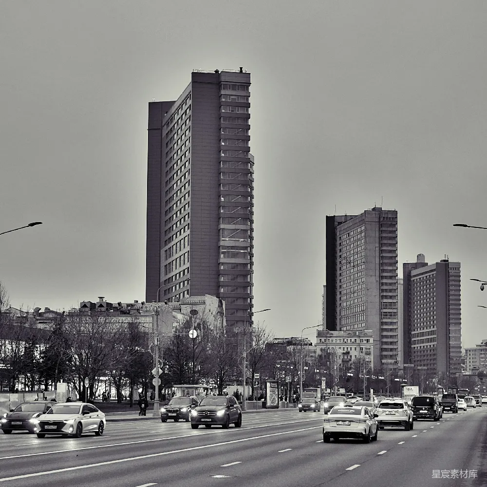 这张黑白照片展示了城市中的高楼大厦与繁忙的街道。画面中，几栋现代化的高楼在晴朗的天空下显得格外突出，街道上车辆川流不息，营造出一种繁忙而有序的城市生活氛围。这幅图像不仅展示了城市的建筑特色，也反映了现代城市中的生活节奏。