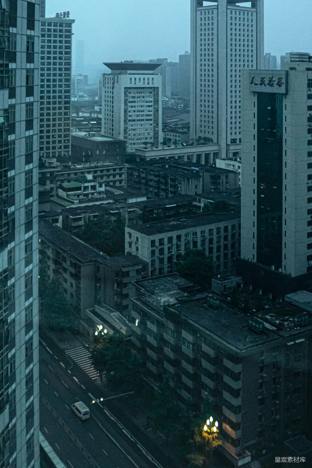 这张图片展示了一个城市的夜晚景象，高楼大厦在昏暗的夜色中显得格外静谧。街道上只有几辆车辆行驶，路边的灯光点缀着城市的宁静。建筑风格多样，既有现代的高楼大厦，也有低矮的居民楼。整体氛围充满了都市的宁静与安详。
