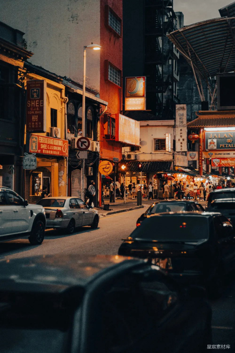 这张图片展示了一条繁忙的城市街道，夕阳的余晖洒在街道上，灯光与招牌的色彩交相辉映，营造出一种热闹而宁静的氛围。街道两旁的建筑物上挂满了各种标志和招牌，人来人往，车辆穿梭，体现了都市生活的繁华与活力。