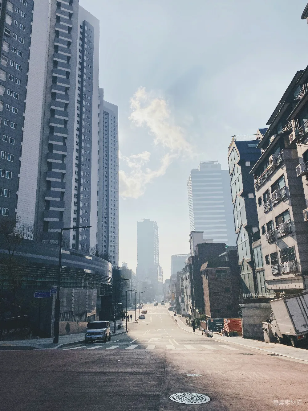 清晨的都市街道，阳光透过云层洒在道路上，建筑群在阳光下显得格外清晰。两侧的高楼大厦与低矮的住宅形成鲜明对比，给人一种宁静而充满活力的感觉。适合用于城市生活、旅游摄影或城市风光的展示。