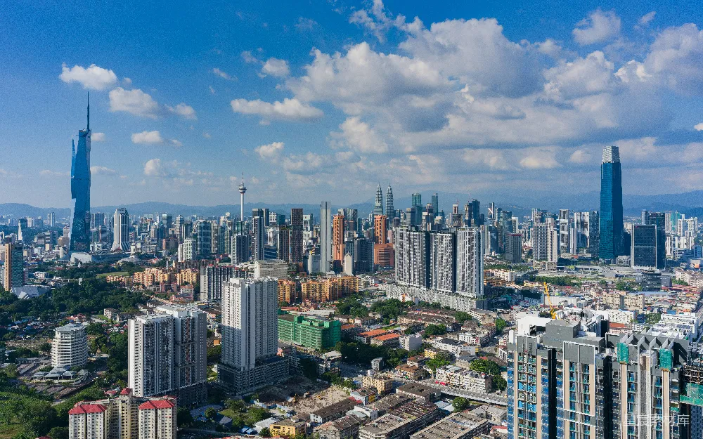 这张图片展示了吉隆坡的城市天际线，展示了现代化的高楼大厦和低矮的住宅楼，背景是蓝天和散布的白云。从空中俯瞰，可以看见城市的布局和建筑的多样性。这张图片适合用作旅游宣传、城市规划展示或现代都市生活背景。