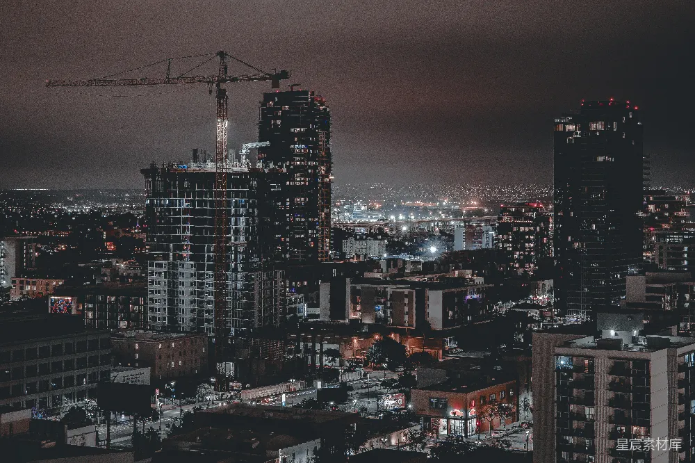 这张图片展示了夜晚的城市景象，画面中心是一座正在建设中的高楼，高耸的起重机在夜色中显得格外醒目。周围的建筑群灯光闪烁，远处的灯光则隐约勾勒出城市的轮廓。整体色调偏暗，营造出一种静谧而略带神秘的氛围，适合用于城市规划、建筑工地或都市生活的主题