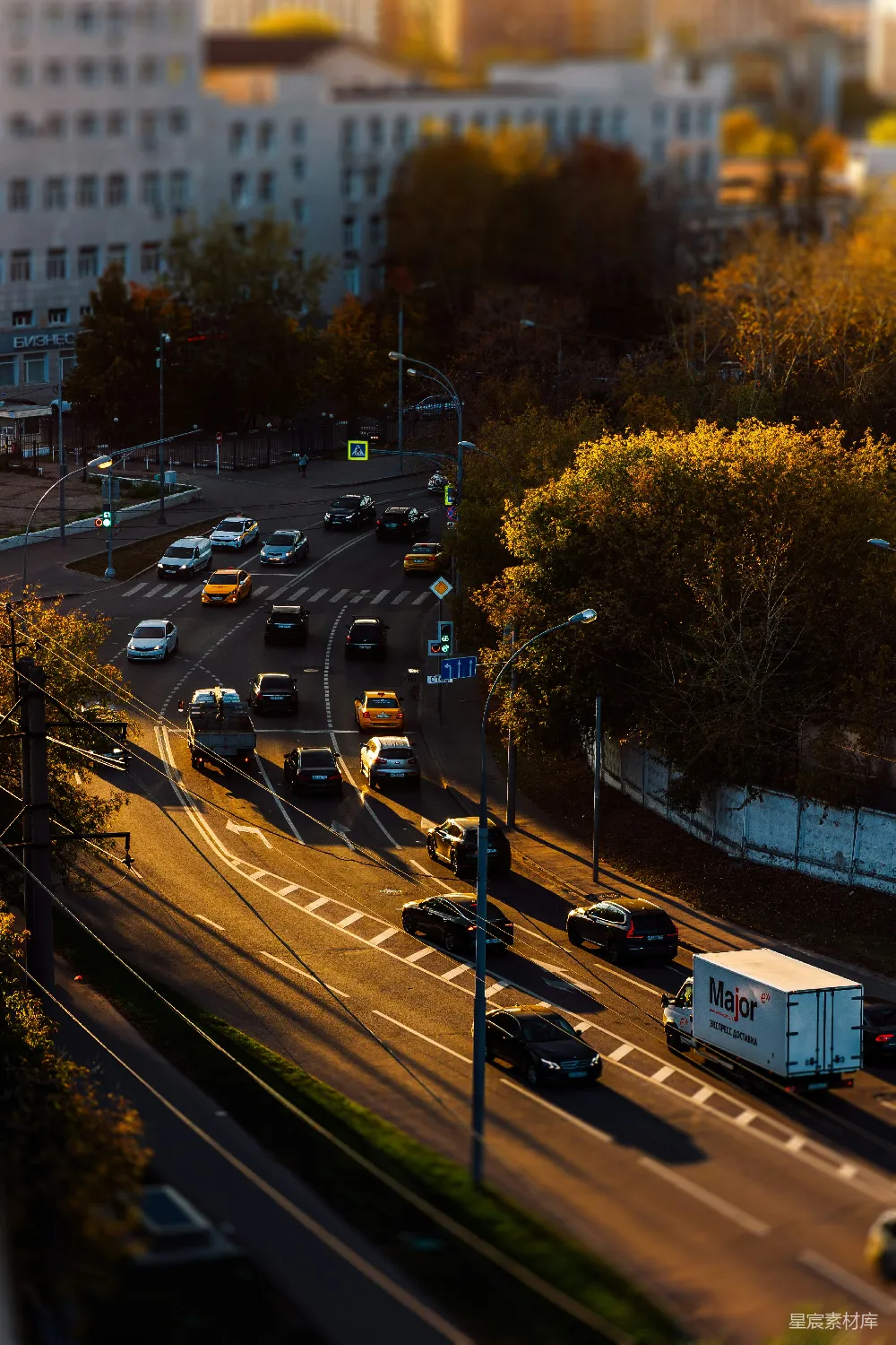 这张图片展示了城市中的繁忙交通场景，阳光洒在道路上，树木呈现出金黄色的秋色，给人一种温暖而宁静的感觉。画面中车辆在十字路口有序行驶，路灯和电线杆错落有致，建筑物背景中透露出都市的繁华。