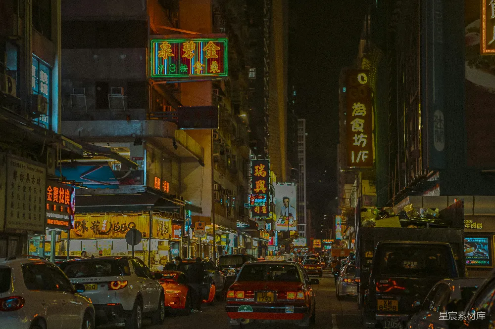 这张图片展示了香港夜晚的繁华街道景象。两侧的建筑上挂满了色彩斑斓的霓虹灯招牌，车流不息，街道两旁的店铺生意兴隆。灯光与车流的动态交织在一起，营造出一种热闹而充满活力的都市氛围。