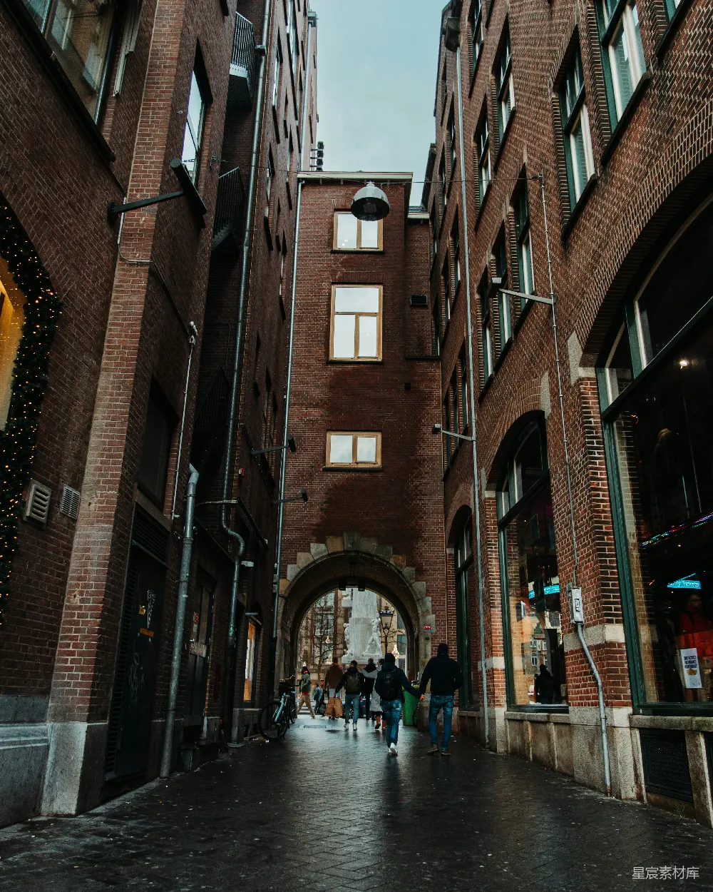 这张图片展示了一条由红砖建筑组成的狭窄巷弄，两旁是高大的建筑物，中间有一座拱门通向更远的地方。巷子里有行人走动，环境显得宁静而富有历史感。适合用来描述城市中的小众景观，或者作为摄影爱好者的灵感来源。