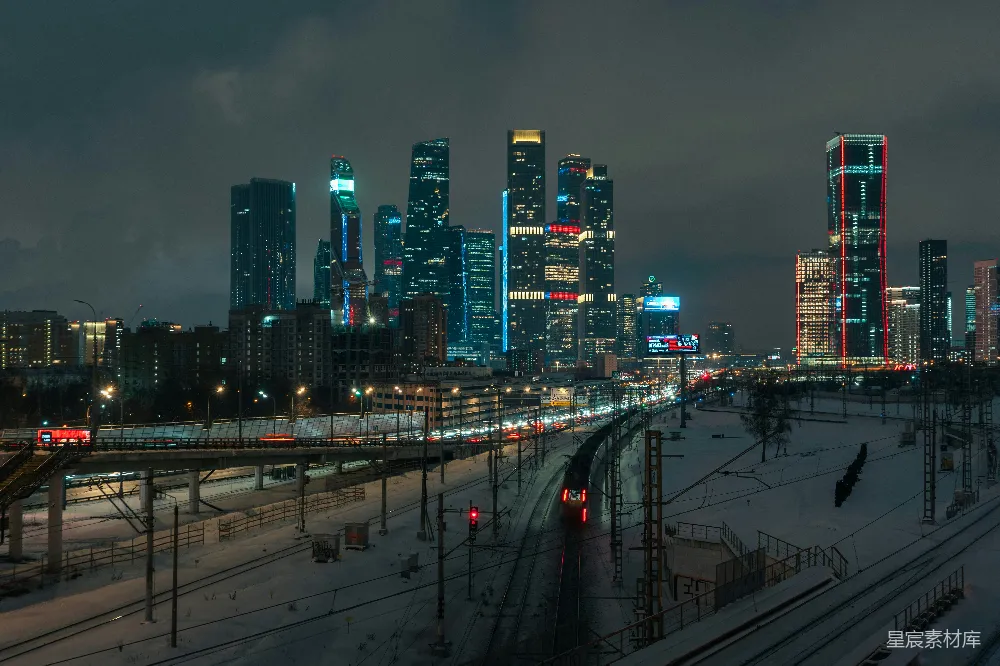这张图片展示了夜晚的城市火车站，背景是灯火辉煌的摩天大楼群。前景中，一列火车正缓缓驶过被积雪覆盖的铁轨，远处的灯光与建筑在黑暗中显得格外醒目，营造出一种静谧而繁华的氛围。适合用于展示城市夜景、火车运输、冬季风光等主题。