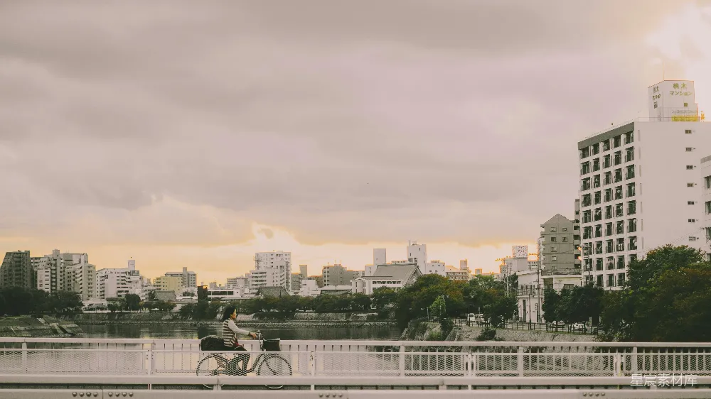 在城市的桥上，一位女士骑着自行车，夕阳的余晖洒在她的身上和远处的建筑上，营造出宁静而美丽的画面。这幅图捕捉了现代都市与自然美景的和谐共存。