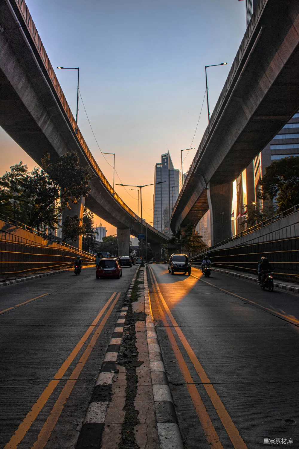 这张图片展示了一条城市中的高架桥，夕阳的余晖洒在桥面上，形成美丽的光影效果。远处可以看到现代化的高楼大厦，道路上有车辆行驶，右侧有树木点缀，整个场景充满了都市的现代感和宁静的氛围。适合用于城市风光、城市生活或旅行相关的场景。