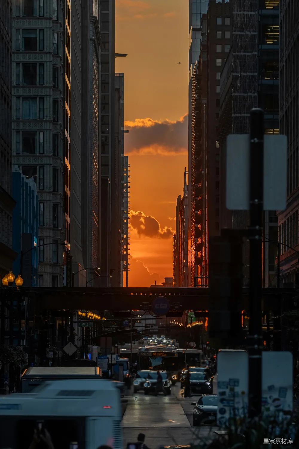 图片展示了一条城市的街道，两旁是高耸的建筑，夕阳的余晖洒在城市上空，形成美丽的渐变色。道路上有行人和车辆，远处的天空中云彩丰富，整个画面充满了宁静与和谐。适合用于城市景观、自然风光、都市生活等场景。