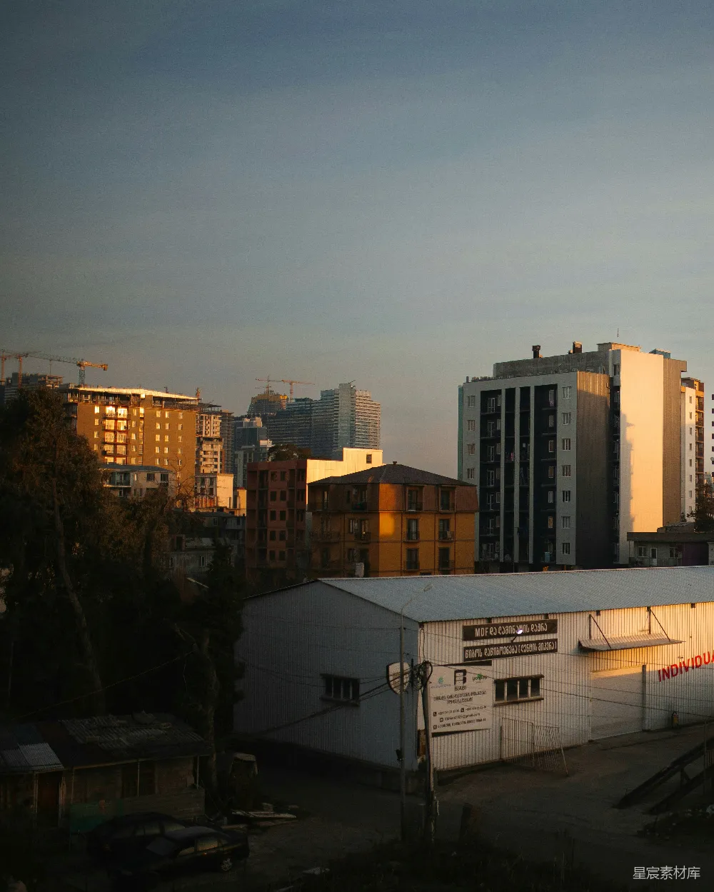 这张图片展示了一个城市的全景，夕阳的余晖洒在建筑物上，形成美丽的光影效果。前景中有一些建筑和工业设施，背景则是正在建设中的高楼大厦，远处的天空呈现出渐变的色彩。整个画面给人一种宁静而富有层次感的感觉，适合用于城市风光、建筑摄影或日常生活场景