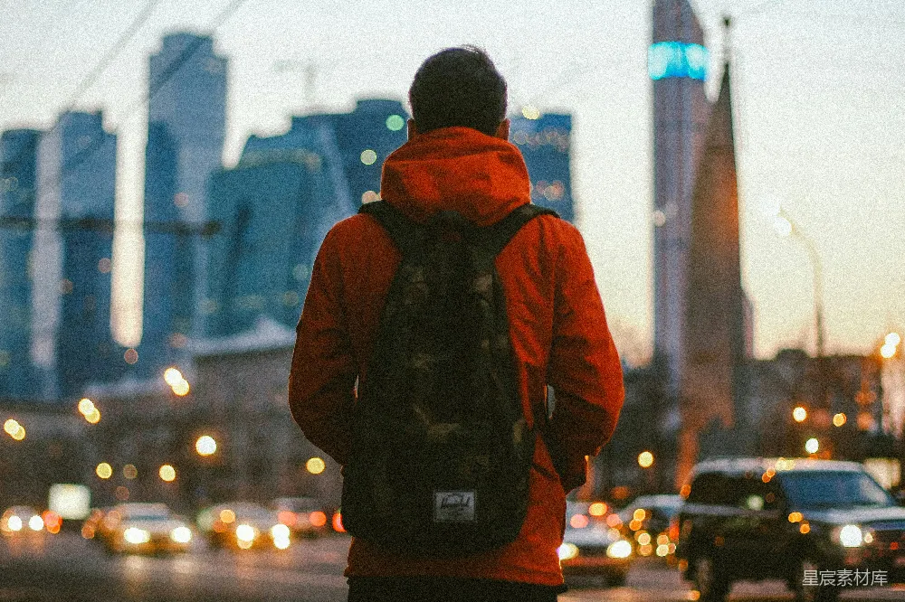 这张图片展示了一个背着背包的旅人在城市夜景中伫立的场景。他身穿红色外套，背对着镜头，面向灯火辉煌的城市天际线。远处高楼大厦的轮廓在夜色中若隐若现，前景中车流的光晕点缀着夜景的美丽。这张图片传达出旅行者探索和发现城市的主题，适合用作旅行、城市