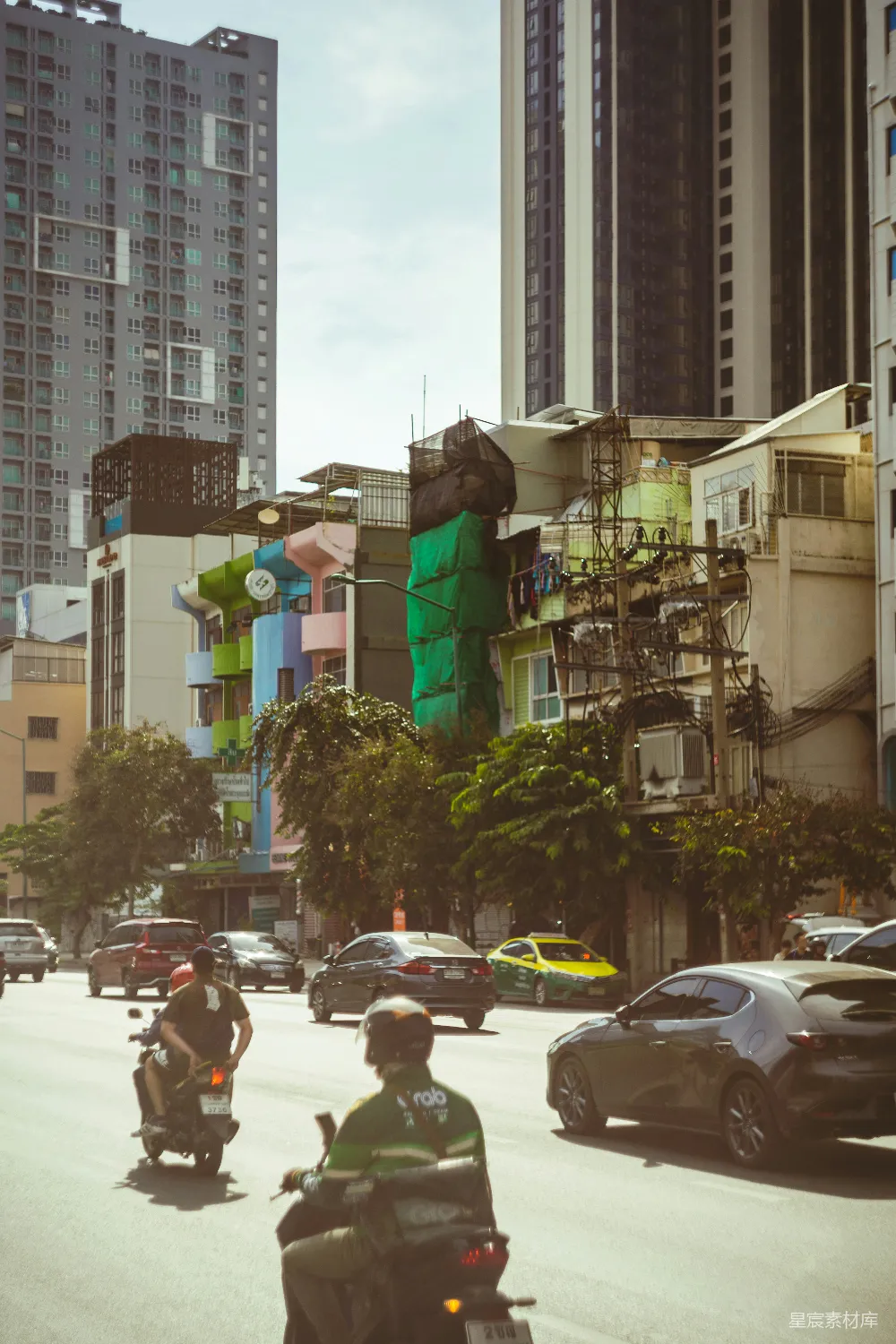 这张图片展示了一个充满活力的城市街道场景。道路上，摩的和汽车交织在一起，形成了繁忙的交通流。背景中的建筑风格多样，既有现代化的高层住宅，也有色彩斑斓的低矮住宅，显示出城市的发展与变化。树木点缀其间，为城市增添了一抹自然的绿意。整体色调温暖，