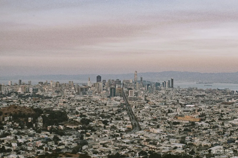 这张图片展示了旧金山的日落美景，城市在夕阳的映照下显得宁静而美丽。远处的天际线与近处的建筑形成了鲜明对比，远处的山峦和海湾也为画面增添了深度。整体色调柔和，给人一种平和与宁静的感觉。适合用于城市风光、日落摄影、旅游宣传等场景。