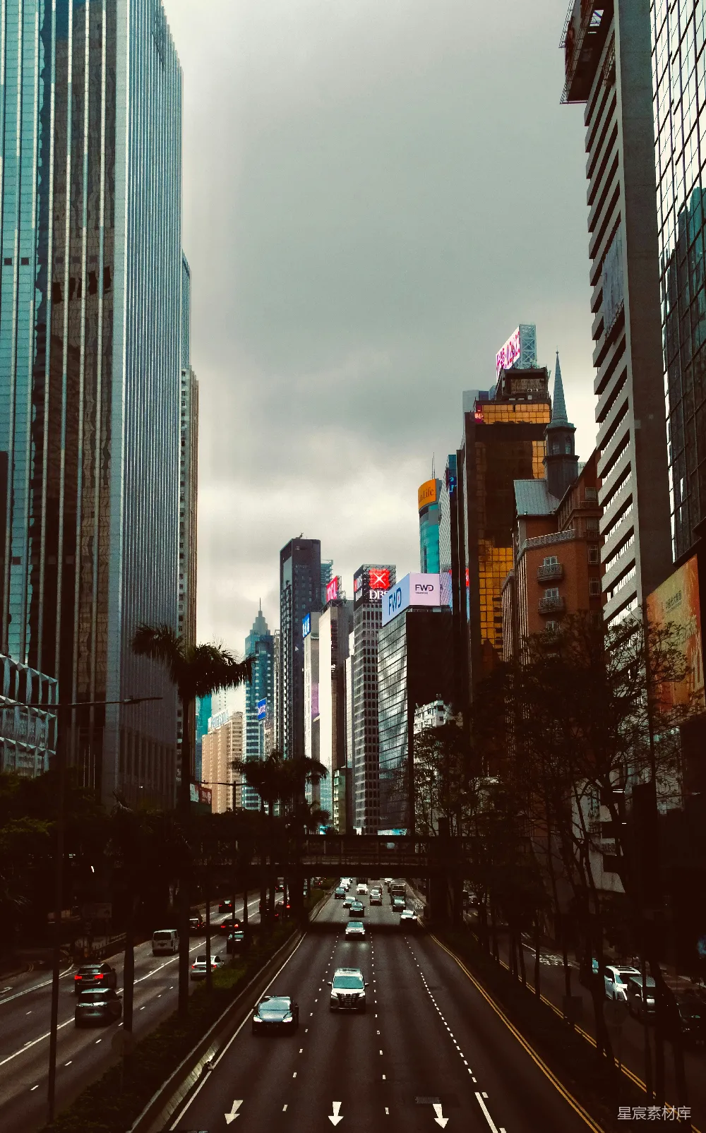 这张图片展示了一条繁忙的城市街道，两侧是高耸的摩天大楼，天空阴沉，给人一种宁静的感觉。街道上有车辆行驶，画面中还有行人和交通信号灯，展现了都市生活的快节奏和宁静的两面。