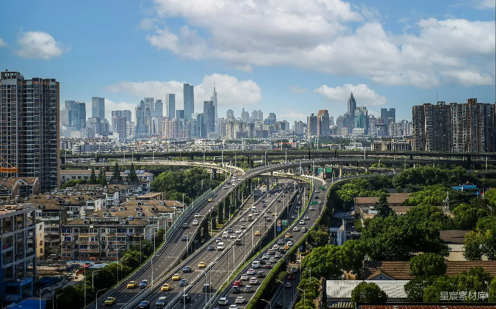 这张图片展示了繁忙的城市高速公路景观，道路两侧是现代化的建筑群和绿色的树木，远处是高耸入云的摩天大楼。天空晴朗，白云点缀，给人一种现代化都市的繁忙与活力。适合用于城市规划、交通管理或现代城市风光的展示。