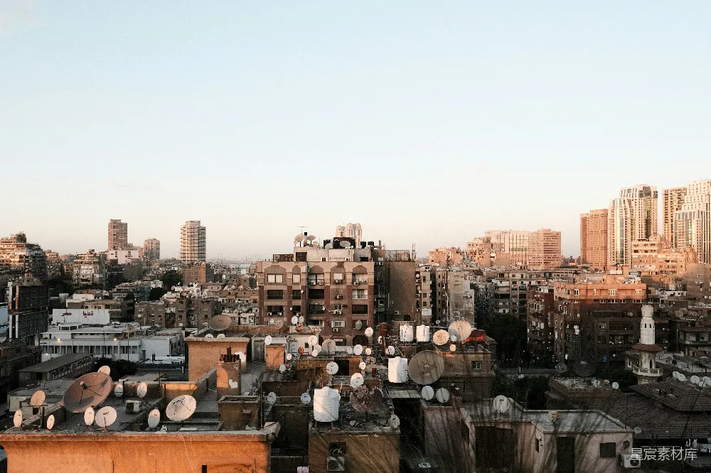 这张图片展示了一个城市天际线在夕阳下的美丽景象。画面中，建筑物的屋顶上布满了卫星天线，天空呈现出温暖的橙色。远处可以看到现代高楼大厦与低矮的住宅楼交织在一起，展现出城市多样化的生活场景。适合用于城市风光、建筑设计或日常生活主题的图片。