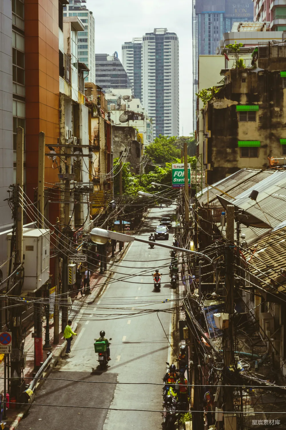 这张图片展示了城市中一条街道的景象，两旁是老旧的建筑和杂乱的电线，背景是现代化的高楼大厦。街道上有人骑摩托车和自行车，电线横跨道路，形成了一种独特的视觉效果。这种场景反映了城市化进程中的复杂性，既有现代的建筑，也有传统的生活方式。