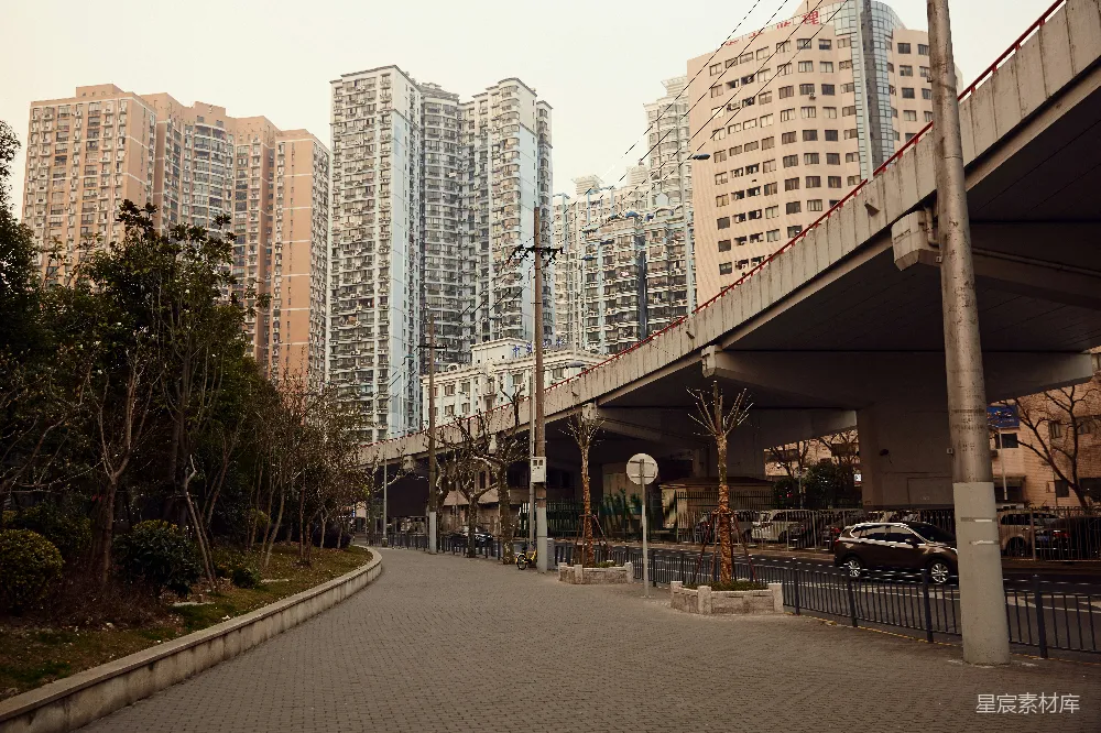 这张图片展示了一条位于城市中的安静小路，两旁是高楼大厦和绿化带。道路宽敞，两旁有路灯和树木，背景中可以看到高耸的住宅楼和桥梁。整体色调偏暖，给人一种宁静的感觉，适合用来表现城市中的静谧时刻，或是城市生活的背景图。