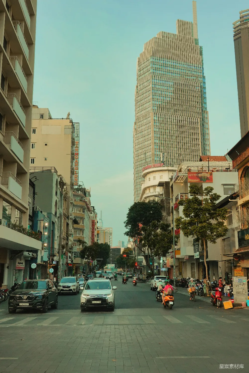 这张图片展示了一条繁忙的都市街道，路两侧是现代化的高楼和低矮的住宅楼，街道上人来人往，车辆穿梭，给人一种繁华和活力的感觉。远处的高楼建筑在夕阳的映衬下显得格外宏伟，而近处的街道则充满了日常生活的气息。这幅画面生动地反映了现代都市的繁忙与都市