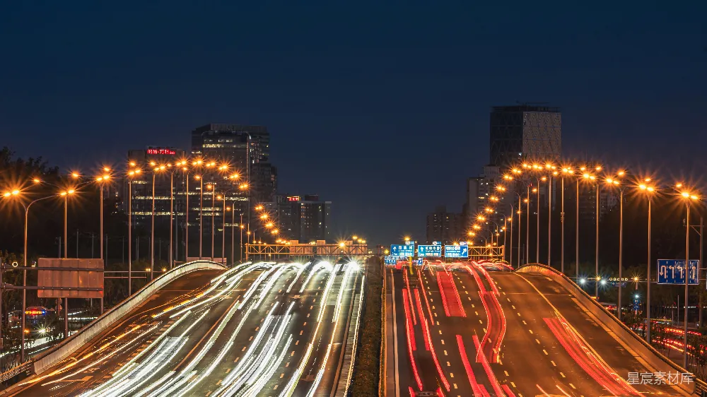 这张图片展示了一条繁忙的高速公路在夜晚的景象。道路两旁的路灯和车流的光带勾勒出清晰的轨迹，背景是高楼大厦和城市的天际线。夜色下的城市充满活力，灯光与黑暗形成了强烈的对比，突显出都市的繁华与快节奏。