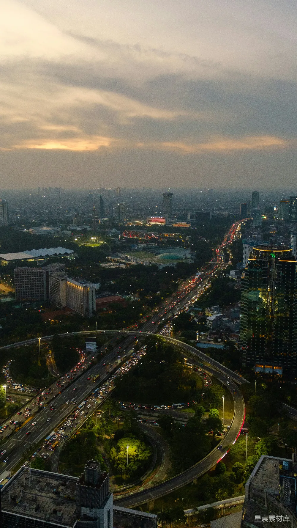 这张图片展示了黄昏时分的城市天际线，天空中布满了柔和的云彩，城市中的高架桥和繁忙的交通线路在灯光的映衬下显得格外引人注目。高楼大厦和公园绿地交织在一起，形成了一幅现代都市的美丽画卷。