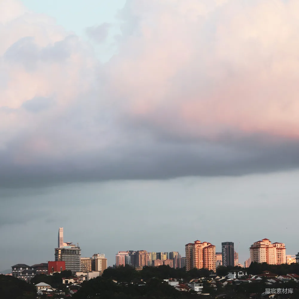 这张图片展示了城市中的建筑群在夕阳照射下的景象。天空中云层厚重，夕阳的余晖为云层染上了柔和的粉红色，与城市的建筑形成了鲜明的对比。远处的树木和近处的建筑在夕阳的余晖中显得宁静而美丽，给人一种平和的感觉。这幅画面不仅展示了城市的景观，也体现了