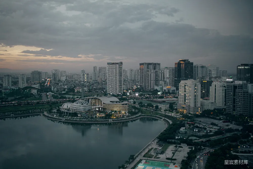 这张图片展示了一座城市的黄昏景象，高楼大厦在昏黄的天空下显得格外醒目。中央有一个圆形的建筑，周围环绕着一个宁静的湖面，湖面上倒映着城市的影子。远处的天空中飘着几朵云彩，给整个画面增添了几分宁静与祥和。适合用作城市风光、建筑摄影或都市生活的主