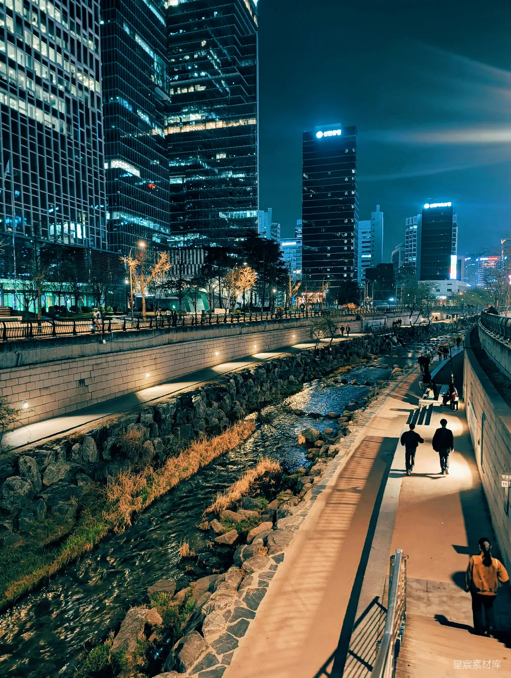 这张图片展示了一个城市的夜晚，河流蜿蜒穿过石墙，两旁是高楼大厦。夜晚的灯光在河面上投射出柔和的光影，形成美丽的倒影。河岸边有行人悠然漫步，享受着夜晚的宁静时光。这张图片体现了都市生活的另一面，既有繁华的现代化建筑，也有自然和人文的和谐共存。