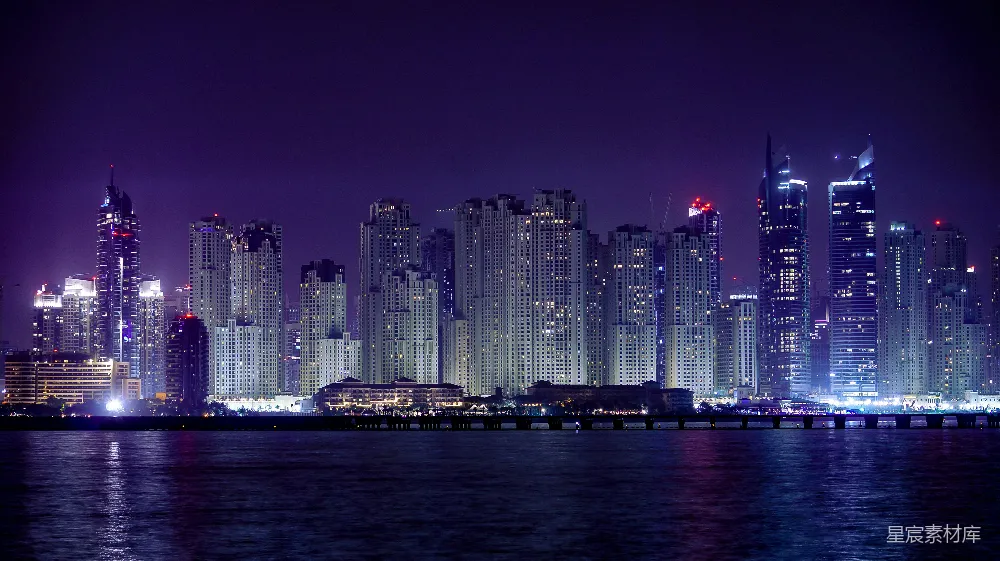 这张图片展示了夜幕下的海滨城市，高楼大厦灯火辉煌，倒映在水面上，形成美丽的光影效果。远处的桥梁连接着城市的两岸，整个画面充满了现代都市的繁华与宁静。适合用于城市风光、旅游宣传或夜景摄影的背景素材。