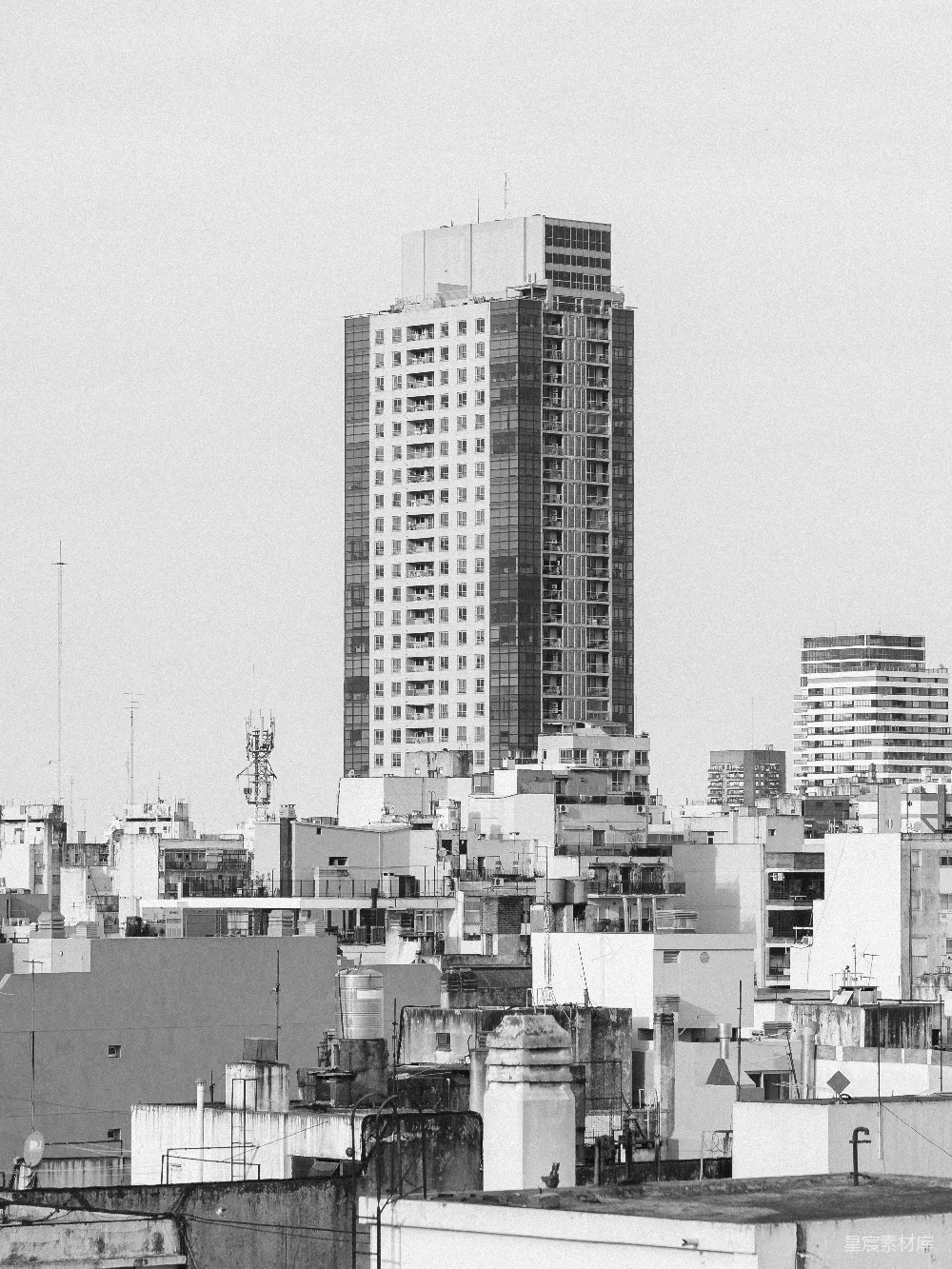 这张黑白照片展示了城市天际线，突出了一座高耸的摩天大楼，周围是低矮的住宅楼。建筑的线条简洁而有力，给人一种现代都市的感觉。适合用于城市景观、建筑设计和现代风格的展示。