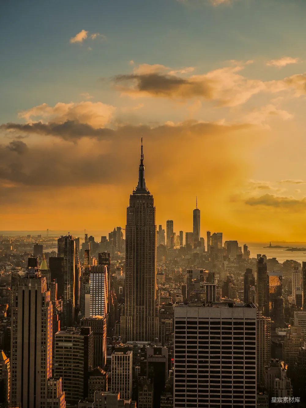 这张图片展示了纽约市天际线的日落景象，中央的帝国大厦在夕阳的余晖中显得格外醒目。城市的建筑在金色的光芒中显得错落有致，背景的天空中飘浮着几朵云彩，为整个画面增添了层次感。这幅图像不仅展示了城市建筑的宏伟，还捕捉到了自然与城市的和谐共存。