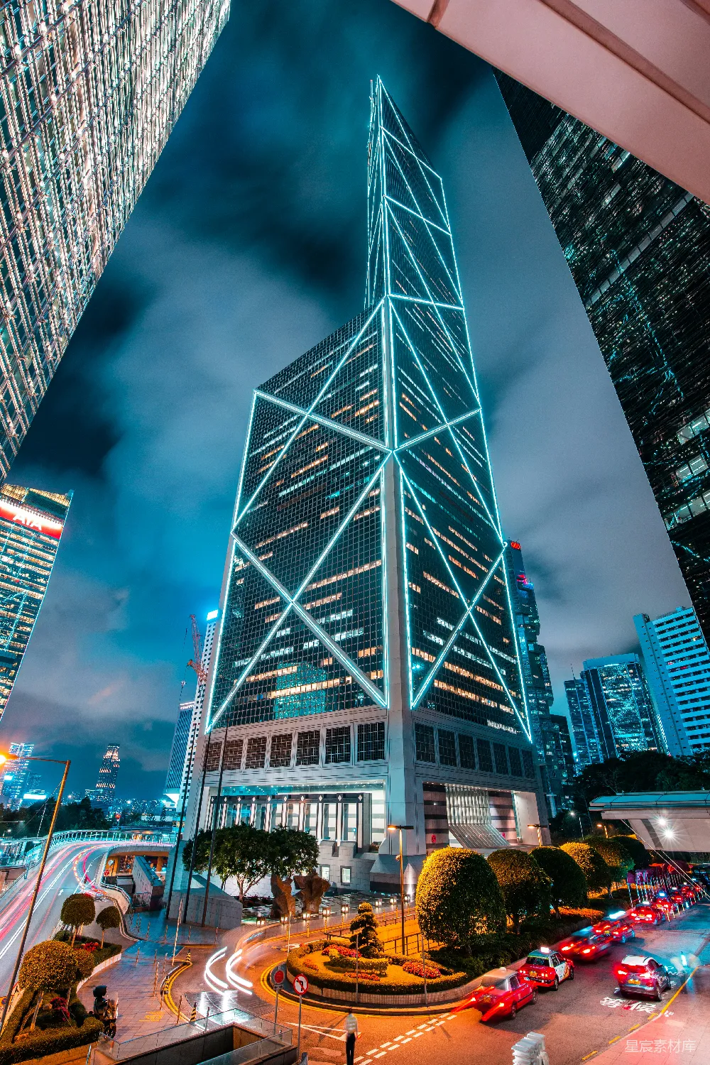 这张图片展示了香港夜晚的高楼大厦，灯光璀璨，现代感十足。建筑的几何线条与灯光效果相得益彰，营造出一种未来感的氛围。街道上的车辆和行人动感十足，增添了城市的活力与动感。适合用于城市风光、现代建筑和都市生活的主题图片。
