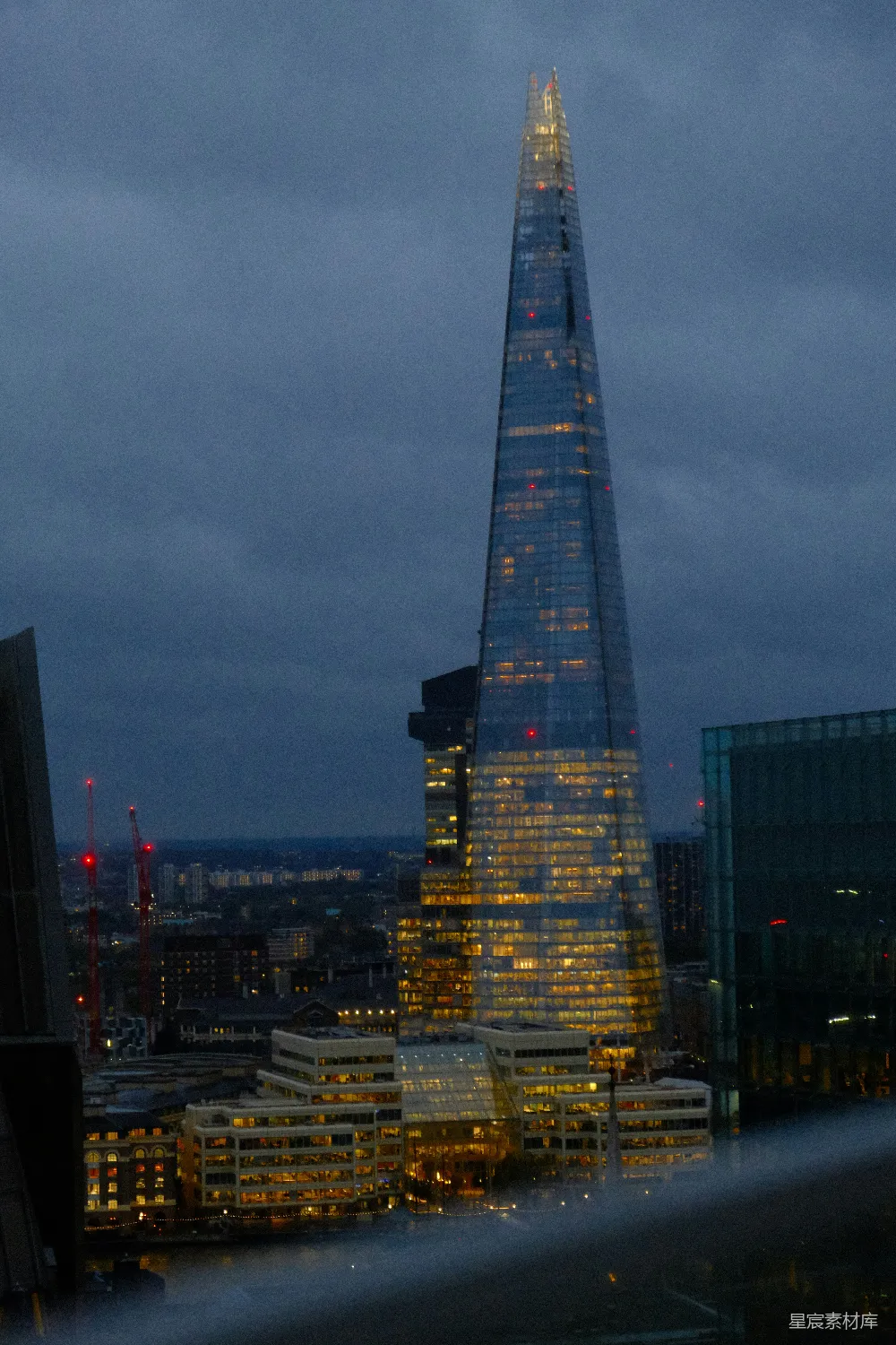 图片展示的是伦敦著名的碎片大厦（The Shard）在黄昏时分的夜景。大厦被灯光照亮，呈现出一种温暖的黄色光芒，与周围昏暗的天空形成鲜明对比。周围的城市景观也隐约可见，展现了伦敦现代化的建筑风格。