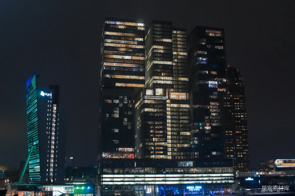 这张图片展示了夜晚灯火辉煌的摩天大楼群，建筑物表面的灯光在黑暗中显得格外醒目。左侧的高楼顶部装饰着蓝色灯光，与右侧的高楼形成鲜明对比。建筑物的玻璃幕墙反射着周围的灯光，营造出一种繁华都市的氛围。适合用于城市夜景、现代建筑或都市风光的背景图片