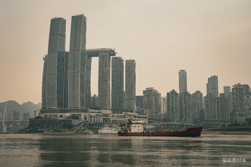 图片展示了一座现代化城市的天际线，高楼林立，建筑风格现代。前景中有一艘红色的货船在江面上行驶，背景则是一片高楼大厦和远处的山脉，天空呈现出淡黄色的色调，给人一种宁静而繁忙的感觉。适合用于城市风光、建筑美学以及工业与自然和谐共存的场景。