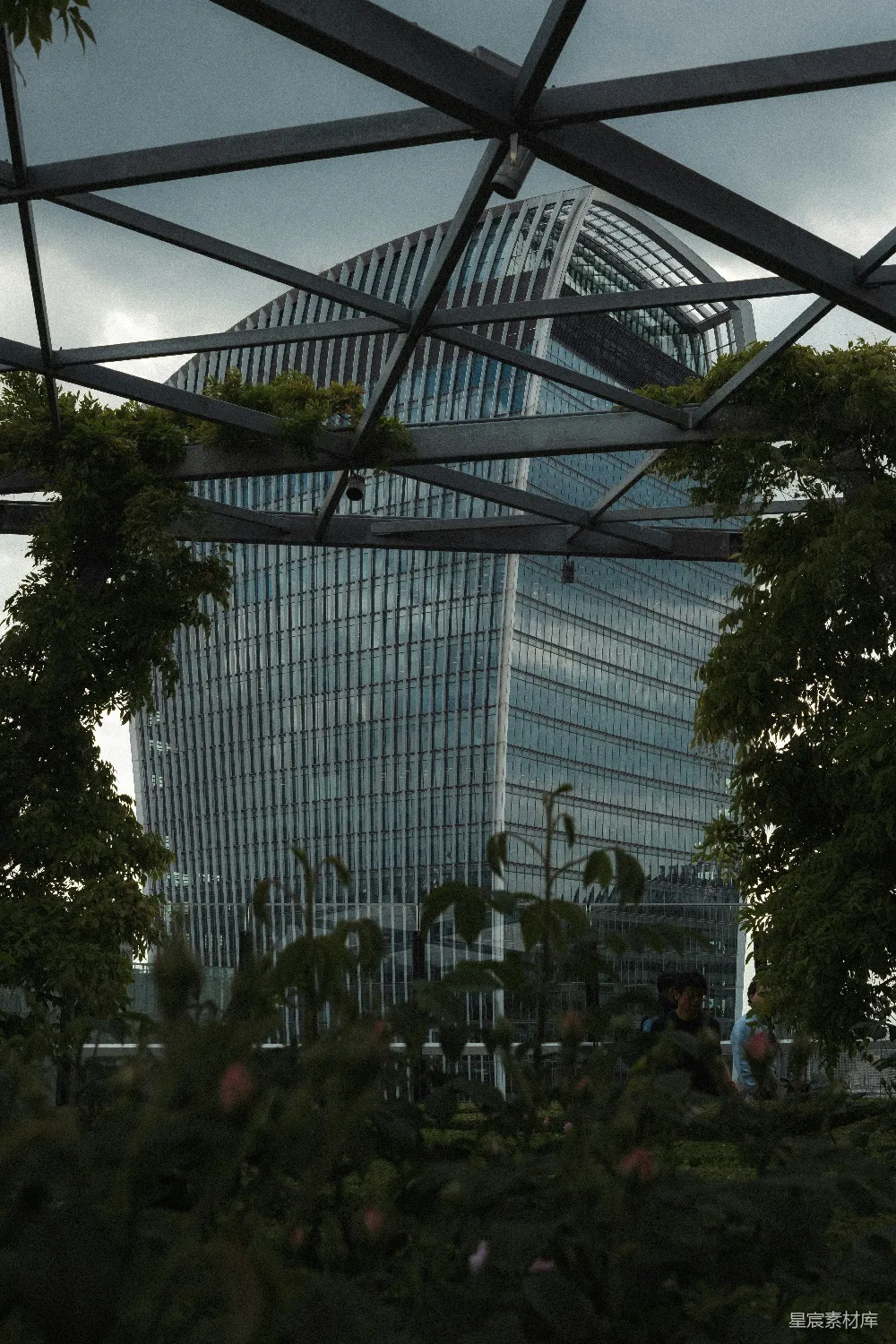 这张图片展示了一座现代高层建筑，其玻璃幕墙反射着周围环境。建筑前的绿色植物和自然景观形成了鲜明对比，体现了城市与自然的和谐共存。整体色调偏冷，给人一种宁静而现代的感觉，适合用于探讨城市绿化、可持续发展或现代建筑美学的主题。
