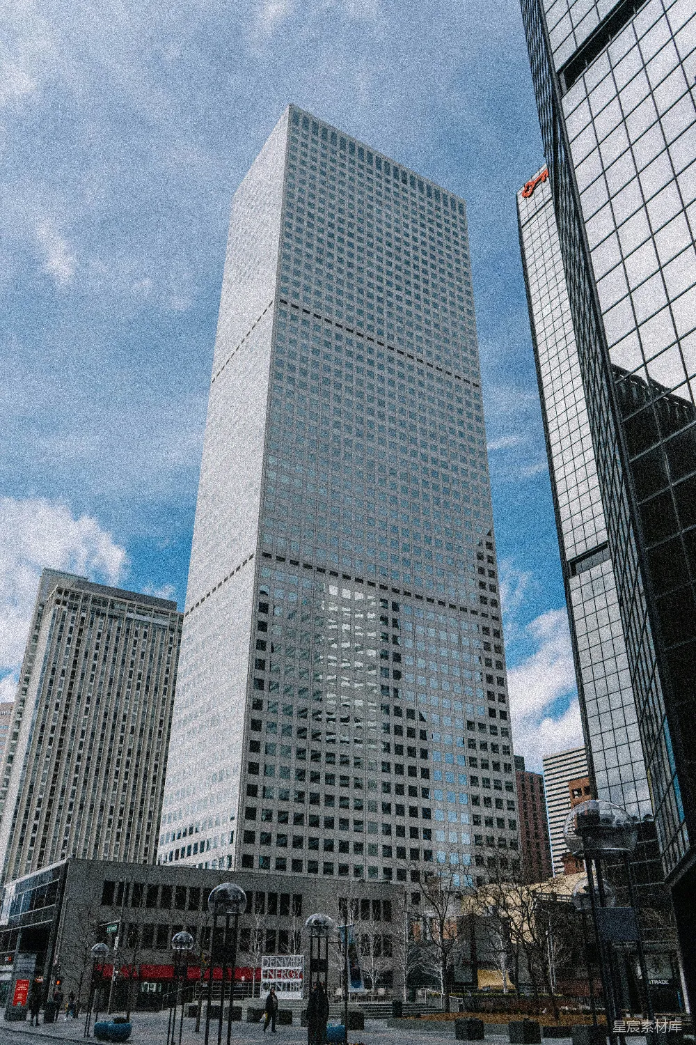 图片展示了一座现代化的高楼建筑，位于城市的中心地带。天空晴朗，几朵白云点缀其中，建筑群线条简洁，玻璃幕墙反射出周围的景致。建筑前有一些行人和交通设施，显示出这是一个繁忙的商业区。整体画面给人一种现代、繁华和充满活力的感觉。