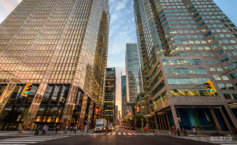 这张图片展示了黄昏时分的城市景象，高楼大厦在夕阳的映照下显得格外辉煌。街道两旁的建筑物反射着夕阳的余晖，街道上有行人和车辆穿梭，整个场景充满了现代都市的活力与繁华。