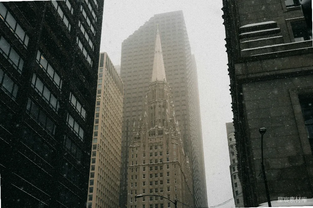 图片中展示了一个雪中的城市景观，雪花在空中飘落，高楼大厦在雪中显得格外庄重。画面中有一座尖塔教堂，与周围的现代建筑形成鲜明对比，营造出一种静谧而神秘的氛围。适合用于展现冬日城市风光或城市建筑的图片素材。