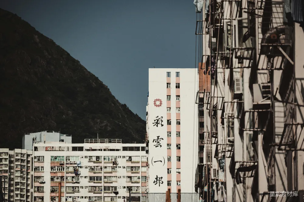 这张图片展示了香港的一处公共住宅区，背景是连绵的山峦，阳光洒在建筑上，营造出一种宁静而略带沧桑的氛围。建筑外墙上醒目地写着“彩云一邨”的字样，窗户上悬挂着晾晒的衣物，透露出居民生活的日常气息。